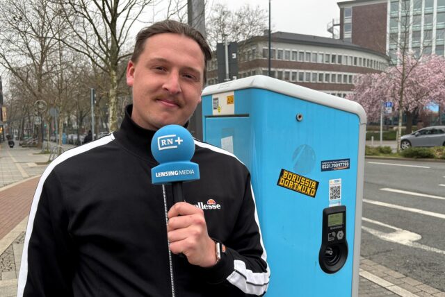 Ruhr Nachrichten-Reporter Tim Hübbertz steht mit einem Mikrofon in der Hand vor einer DEW-Ladesäule in Dortmund.