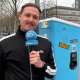 Ruhr Nachrichten-Reporter Tim Hübbertz steht mit einem Mikrofon in der Hand vor einer DEW-Ladesäule in Dortmund.
