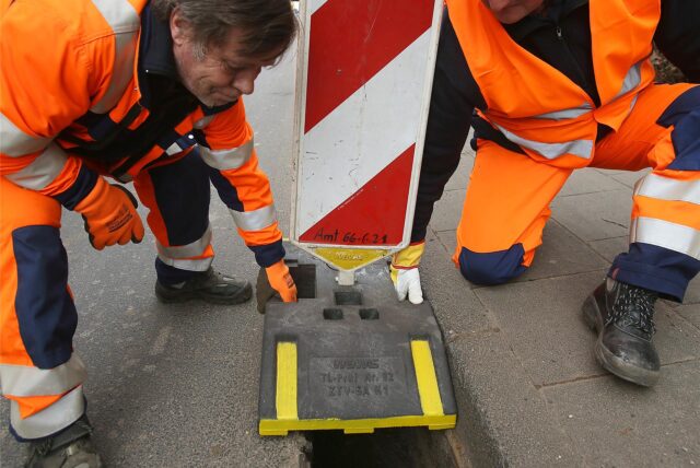 Mitarbeiter einer Straßenmeisterei decken einen Abfluss am Straßenrand mit einer Warnbake ab.