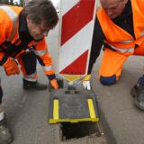 Mitarbeiter einer Straßenmeisterei decken einen Abfluss am Straßenrand mit einer Warnbake ab.