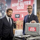 Joko Winterscheidt und Klaas Heufer-Umlauf vor einem Plakat zur Live-Quizshow „Ein sehr gutes Quiz (mit hoher Gewinnsumme)“ und mit einem Geldkoffer, der die Aufschrift „Hohe Gewinnsumme zahlt ProSieben“ trägt