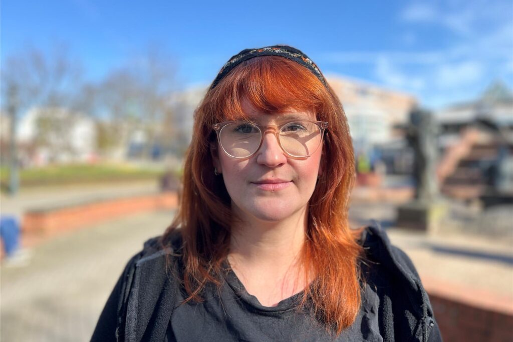 Eine rothaarige Frau mit Brille schaut in die Kamera. Im Hintergrund unscharf zu erahnen: der Stadtgarten in Dortmund. Eine rothaarige Frau mit Brille schaut in die Kamera. Im Hintergrund unscharf zu erahnen: der Stadtgarten in Dortmund.