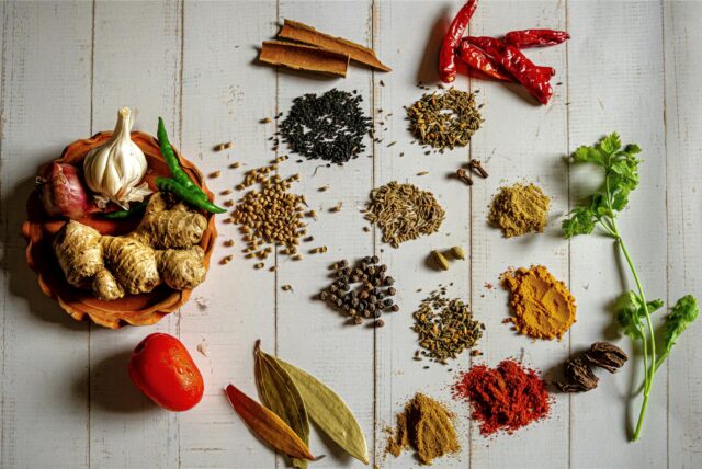 Auf einem weißen Holztisch liegen verschiedene Gewürze – unter anderem Zimt, Paprika, Pfeffer und Kümmel.