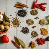 Auf einem weißen Holztisch liegen verschiedene Gewürze – unter anderem Zimt, Paprika, Pfeffer und Kümmel.