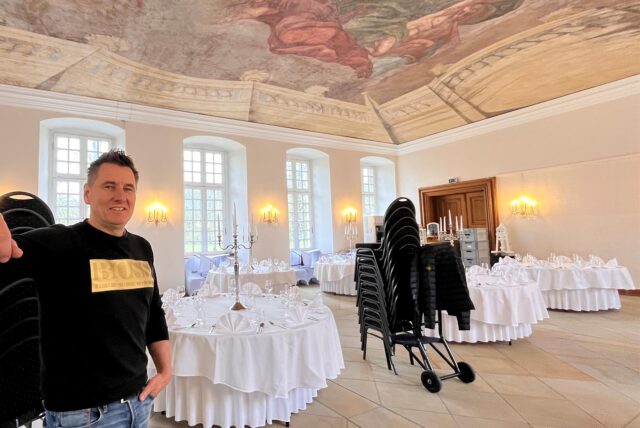 Markus Liptow, Gastronom im Schloss Herten, steht im Speisesaal. Hinter ihm in weiß eingedeckte Tische und ein Stuhlwagen für den Aufbau der Bestuhlung.