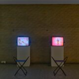 Zwei Fernsehmonitore mit Videos darauf, vor jedem der Bildschirme steht ein Sitzhocker.