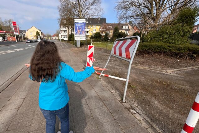 Westerholterin mit blauer Jacke und langen Haaren steht mit der Geste des Unverständnisses vor dem verbogenen Schild.