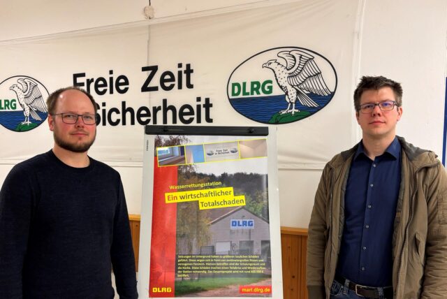 Martin Gertz und Stephan Spiecker stehen im Vereinsheim der Marler DLRG Ortsgruppe. Zwischen sich steht ein Plakat mit der Aufschrift, „Wasserrettungsstation ein wirtschaftlicher Totalschaden.“ Im Hintergrund ist ein Plakat mit der Aufschrift „Freie Zeit für Sicherheit“ zu sehen. Es wurde vor einen breiten Riss in der Wand gehängt, der durch das Plakat erkennbar ist.  Der Riss wurde an einer Stelle mit Spachtelmasse abegdeckt.