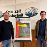 Martin Gertz und Stephan Spiecker stehen im Vereinsheim der Marler DLRG Ortsgruppe. Zwischen sich steht ein Plakat mit der Aufschrift, „Wasserrettungsstation ein wirtschaftlicher Totalschaden.“ Im Hintergrund ist ein Plakat mit der Aufschrift „Freie Zeit für Sicherheit“ zu sehen. Es wurde vor einen breiten Riss in der Wand gehängt, der durch das Plakat erkennbar ist.  Der Riss wurde an einer Stelle mit Spachtelmasse abegdeckt.