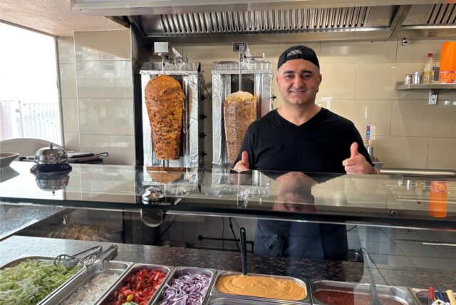 Ein Mann steht hinter einer Theke mit Salat und Saucen. Hinter ihm zwei Dönerspieße.