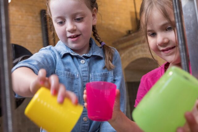 Zu sehen sind zwei Kinder, die mit bunten Plastikbechern spielen. Hinter ihnen ist ein großes Backsteingebäude