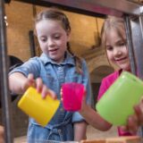 Zu sehen sind zwei Kinder, die mit bunten Plastikbechern spielen. Hinter ihnen ist ein großes Backsteingebäude