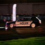 Ein BMW Dreier Touring aus den frühen 2000ern ist vor dem Hanger des Flugplatzes Loemühle zu sehen. Er driftet vor einer Absperrung mit Zuschauern. Auf der Seite des schwarzen und weißen Auots steht „Police“. Vom Vorderrad in Richtung Fahrertür geht ein aufgemalter Mund.
