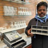 Neben einer, mit weißem Staub bedeckten, Kasse und zwei Ablagen mit Gläsern, ebenfalls mit weißem Staub bedeckt, steht ein Mann mit einem leeren Besteckbehälter in der Hand. Dieser ist auch von weißem Staub bedeckt.