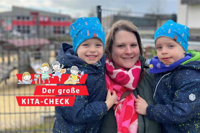Eine Mutter hat ihre zwei Jungen im Kindergartenalter auf dem Arm. Im Hintergrund unscharf zu erkennen: der Spielplatz einer Kita hinter einem Zaun.