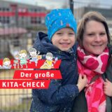 Eine Mutter hat ihre zwei Jungen im Kindergartenalter auf dem Arm. Im Hintergrund unscharf zu erkennen: der Spielplatz einer Kita hinter einem Zaun.