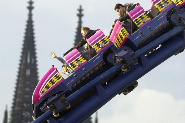 Besucher sitzen auf der Deutzer Kirmes in Köln in einer Achterbahn, während im Hintergrund die Türme des Kölner Doms zu sehen sind.