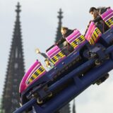 Besucher sitzen auf der Deutzer Kirmes in Köln in einer Achterbahn, während im Hintergrund die Türme des Kölner Doms zu sehen sind.