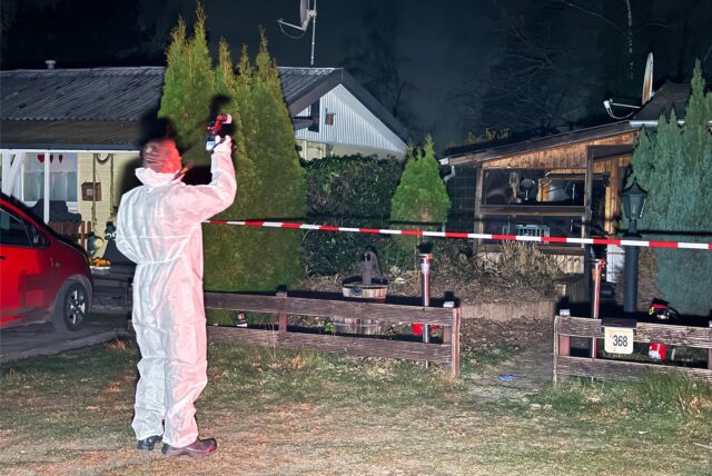 Mann im Schutzanzug macht Fotos an einer ausgebrannten Hütte.