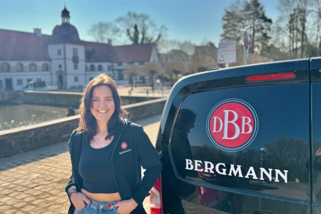 Anna Pusch, Pressesprecherin der Bergmann-Brauerei, steht vor dem Wasserschloss Haus Rodenberg in Dortmund-Aplerbeck. Neben ihr steht ein Transporter, auf dem das Bergmann-Logo zu sehen ist.