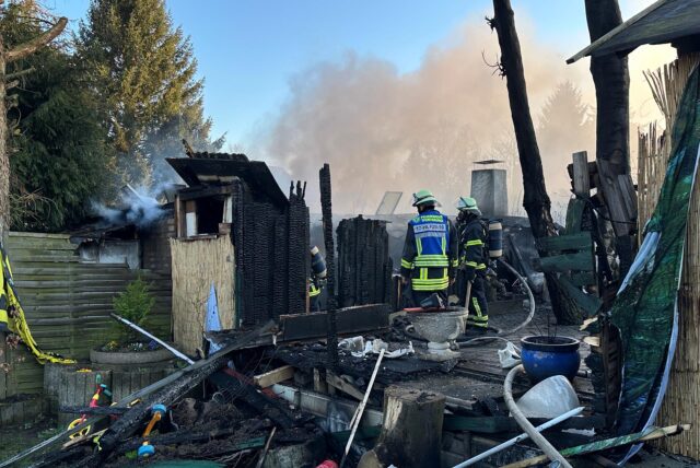 Einsatzkräfte der Feuerwehr stehen zwischen den heruntergebrannten Überresten von Gartenlauben in Dortmund-Menglinghausen.