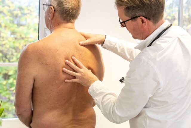 Ein oberkörperfreier Mann mittleren Alters steht mit dem Rücken zum Arzt, der in weißem Kittel den Rücken untersucht.