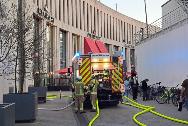 Ein Löschfahrzeug der Feuerwehr steht vor dem Palais Vest in Recklinghausen. Gelbe Schläuche liegen auf dem Boden. Neben Passanten sind auch zwei Feuerwehrleute in Montur zu sehen.