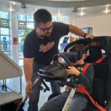 Der junge Paul sitzt im Glashaus Herten in einem Fahrsimulator, während ihm Patrick Becker die Funktionen erklärt.