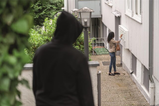 Ein schwarz gekleideter Mann lauert einer Frau vor einem Haus auf und beobachtet sie.