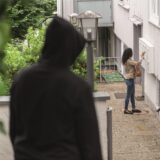 Ein schwarz gekleideter Mann lauert einer Frau vor einem Haus auf und beobachtet sie.