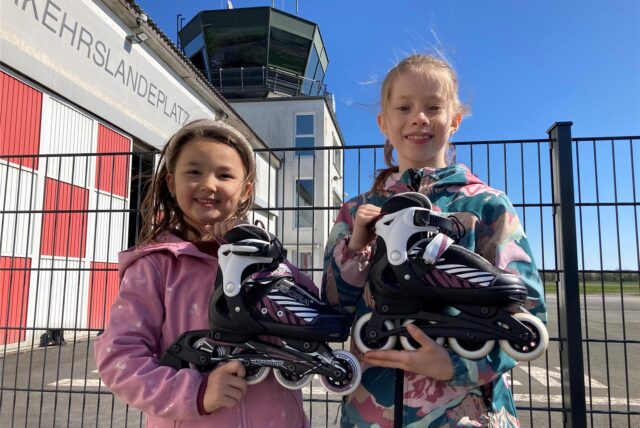 Leni Ciolek (9 Jahre, rechts) und Hailey Huynh halten Inlineskates in den Händen, hinter ihnen ist der Tower des Flugplatzes Loemühle.