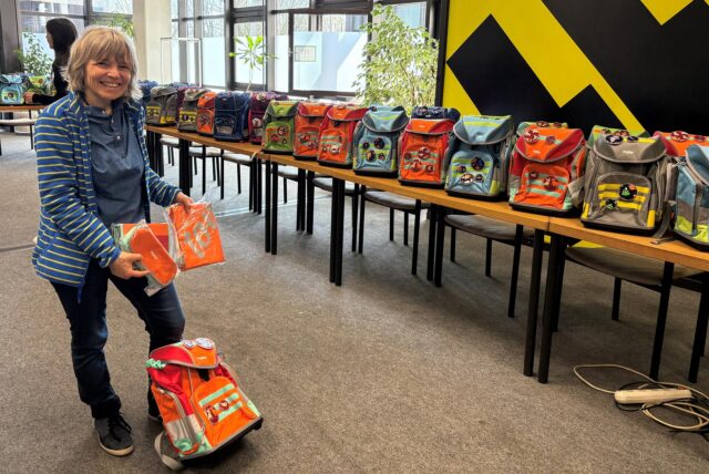 Eine Frau steht im Vordergrund und hält einen Turnbeutel, ein Federmäppchen und ein Etui vor sich. Zu ihren Füßen steht ein Tornister. Im Hintergrund sind Tische mit weiteren Schulranzen zu sehen.