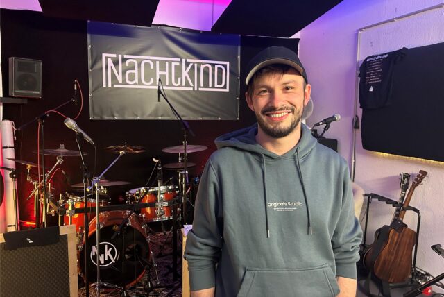 Timo Schmitt steht im Proberaum, hinter ihm Instrumente und ein Banner mit der Aufschrift „Nachtkind“.
