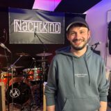 Timo Schmitt steht im Proberaum, hinter ihm Instrumente und ein Banner mit der Aufschrift „Nachtkind“.