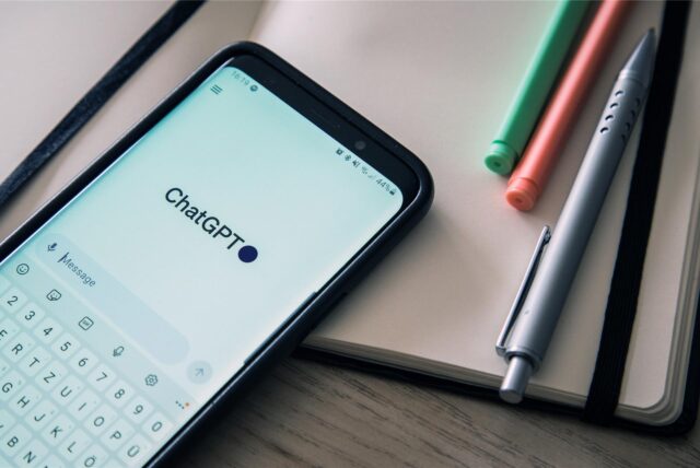 Auf einem Tisch liegen ein Smartphone, auf dem ChatGPT geöffnet ist, ein Notizbuch und Stifte.