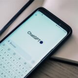 Auf einem Tisch liegen ein Smartphone, auf dem ChatGPT geöffnet ist, ein Notizbuch und Stifte.
