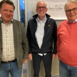 Drei Männer mittleren Alters stehen nebeneinander und lächeln in die Kamera. Sie tragen Freizeitkleidung und Brillen.