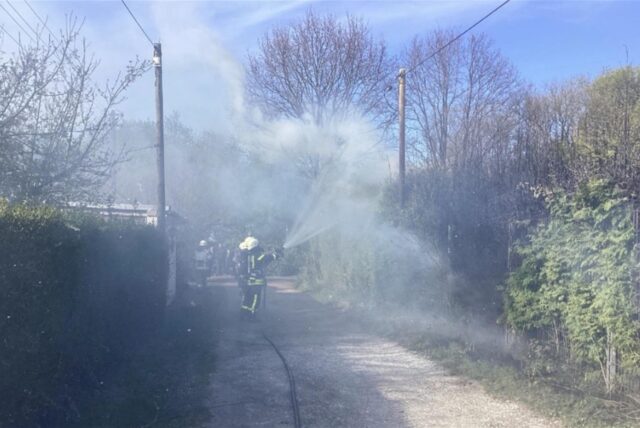 Rauch steigt aus einer Hecke empor. Davor stehen Feuerwehrleute unter Atemschutz und halten mit C-Rohren auf das Feuer.