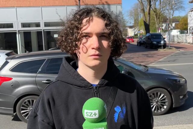 Ein junger Mann mit lockigen Haaren guckt in die Kamera und hält ein Mikrofon in der Hand.