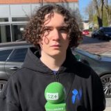 Ein junger Mann mit lockigen Haaren guckt in die Kamera und hält ein Mikrofon in der Hand.