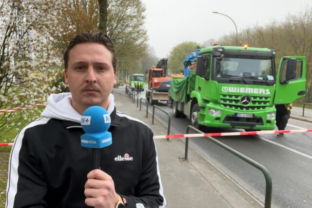 Reporter Tim Hübbertz von den Ruhr Nachrichten steht mit einem Mikrofon vor der Kamera. Im Hintergrund sind mehrere Arbeitsfahrzeuge zu sehen, darunter ein grüner LKW der Firma Wiemers.