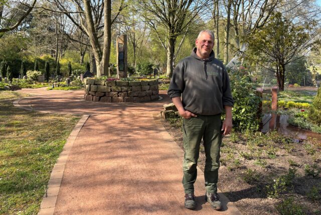 Gärtnermeister Andreas Gehron steht auf dem Hauptfriedhof in Marl. Im Hintergrund sind Anpflanzungen und Gräber und eine laufende Sprinkleranlage zu sehen.