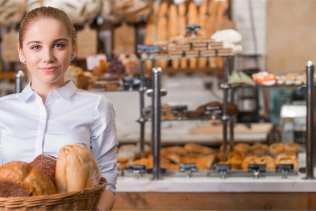 Bäckerin vor einem Ofen hält ein Blech mit Baguettes.