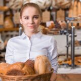 Bäckerin vor einem Ofen hält ein Blech mit Baguettes.
