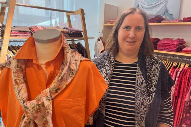 Eine Frau steht in einem Bekleidungsgeschäft neben einer Schaufensterpuppe und Regalen und Stangen voller Kleider.