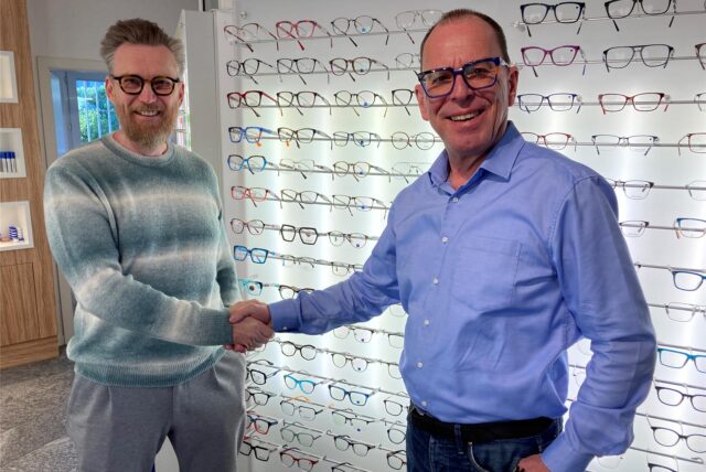 Zwei Marler Optiker reichen sich von einer Wand voller Brillen die Hand.