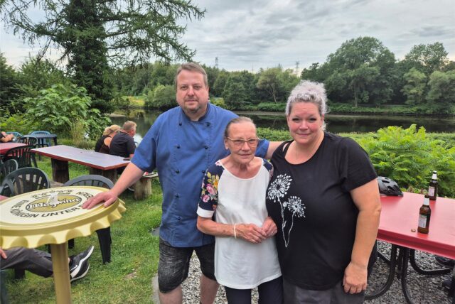 Drei Personen posieren für ein Foto im Freien, mit einem Fluss und Bäumen im Hintergrund. Im Vordergrund sind Tische und Stühle zu sehen, was auf einen Garten oder eine Terrasse hindeutet.