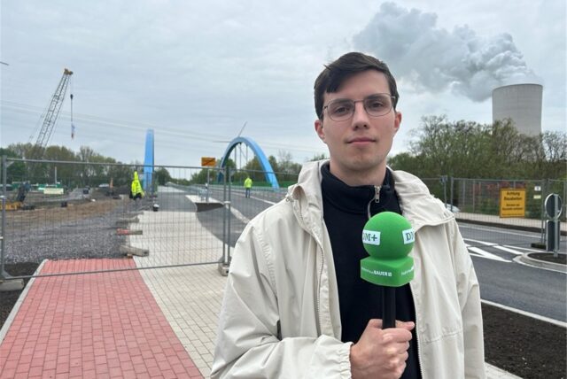 Das Bild zeigt eine neu gebaute Brücke mit blauen Bögen, die einen Fluss oder Kanal überspannt. Davor steht ein Reporter.