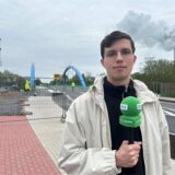 Das Bild zeigt eine neu gebaute Brücke mit blauen Bögen, die einen Fluss oder Kanal überspannt. Davor steht ein Reporter.