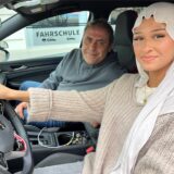 Eine junge Frau sitzt am Steuer eines Autos. Auf dem Beifahrersitz sitzt ein Mann mit langen Haaren. Beide lächeln in die Kamera.
